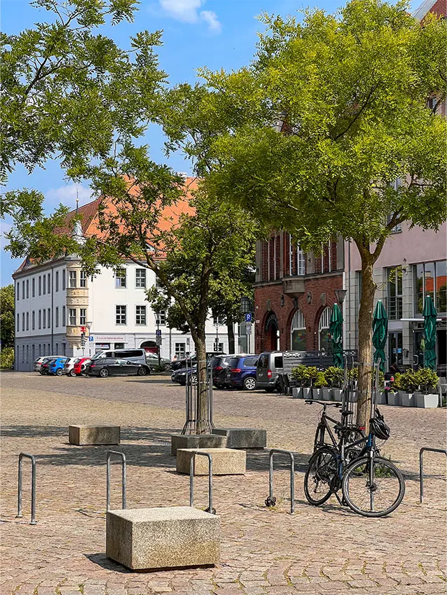 WOWI-FFO-Stadtteil-Zentrum: Platz mit Bäumen, Bänken, Fahrradständern und Gebäuden im Hintergrund. Autos parken.