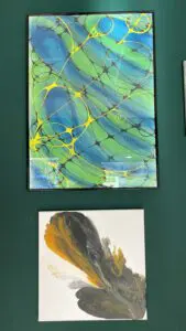 Wander-Atelier der Talente: Gerahmtes abstraktes Kunstwerk in Blau, Grün und Gelb. Ein weiteres Kunstwerk darunter.