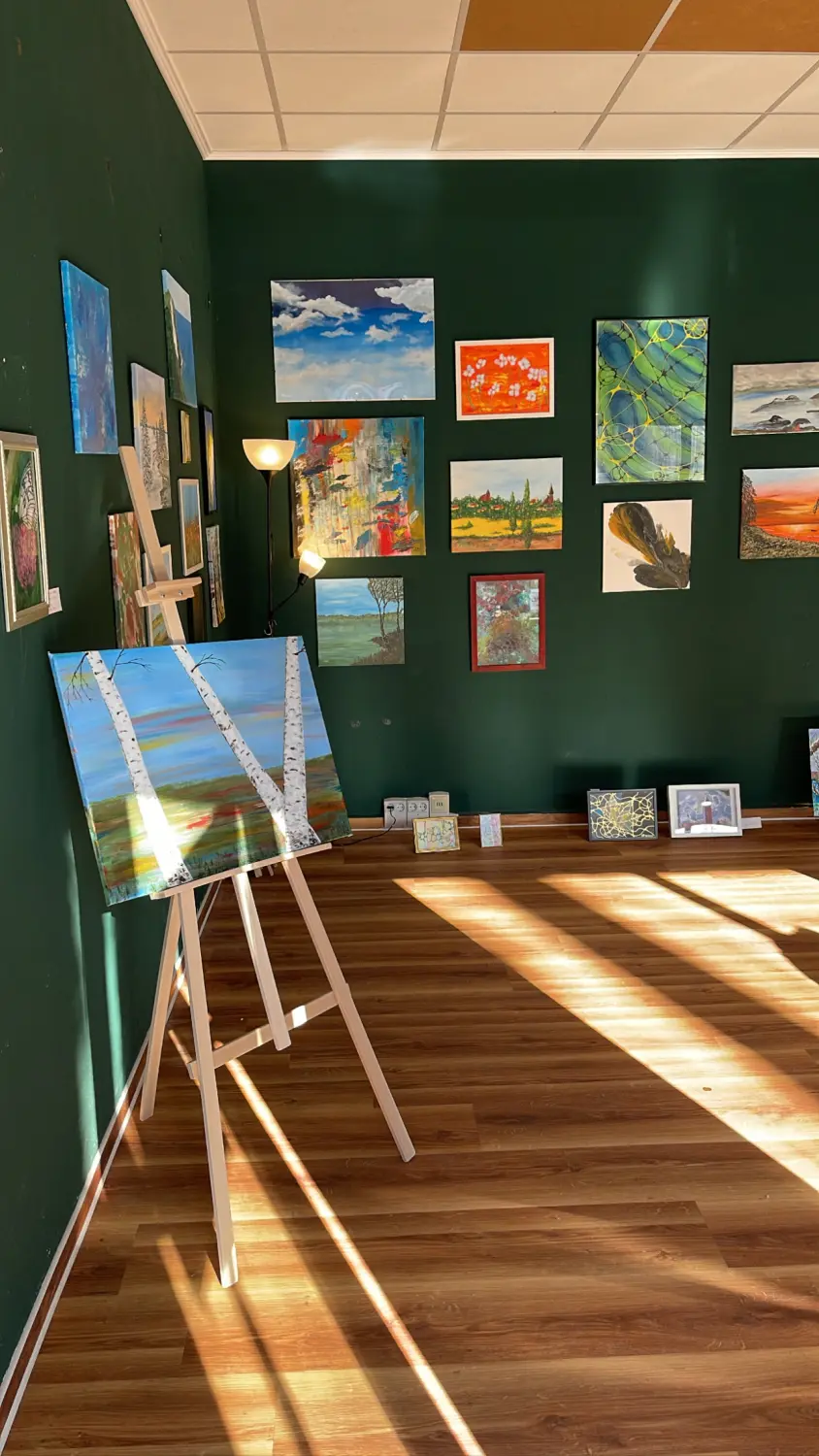 Wander-Atelier der Talente: Gemäldeausstellung mit Birkenmotiv auf Staffelei, umgeben von weiteren Kunstwerken an grüner Wand.