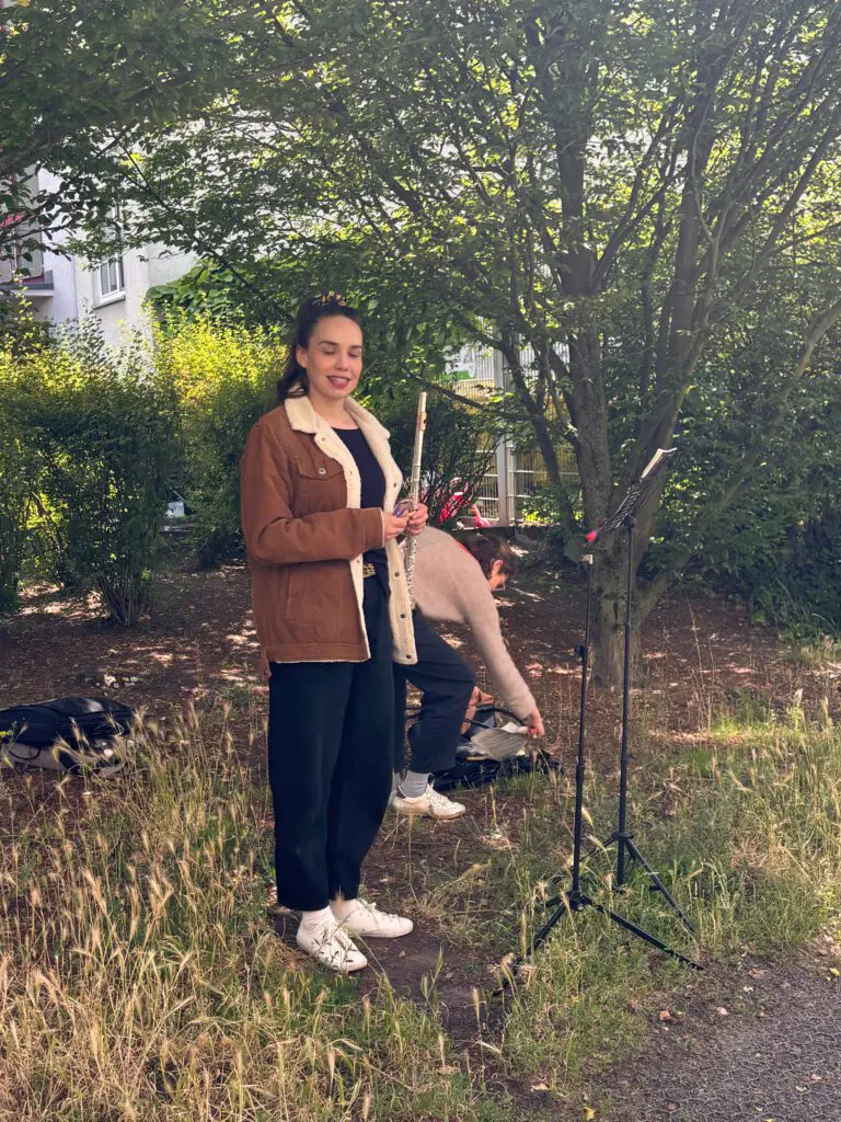 Eine Flötistin in brauner Jacke hält bei Treppenhausmusik 2027 vor einem Notenständer inne, während eine Person sich bückt