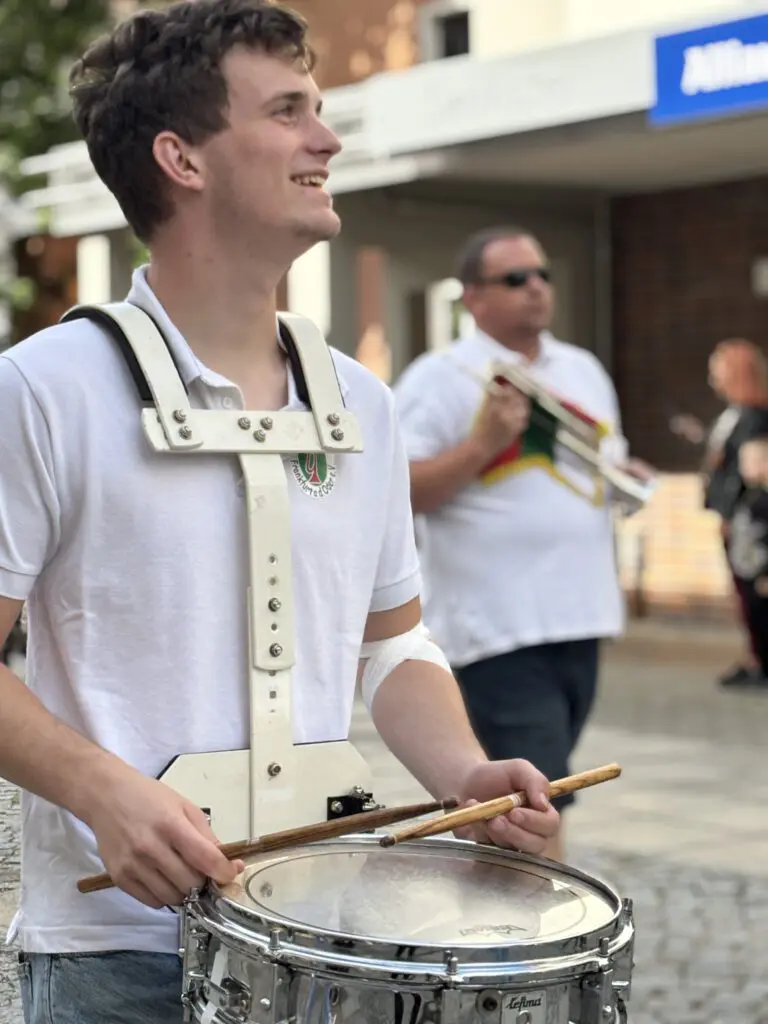Ein laechelnder Musiker mit weissem Poloshirt traegt bei Treppenhausmusik 2027 eine Snare Drum vor einem weiteren Spieler