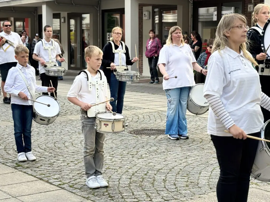 Mehrere Menschen in weissen Oberteilen trommeln bei Treppenhausmusik 2027 auf dem gepflasterten Boden vor einem Gebaeude