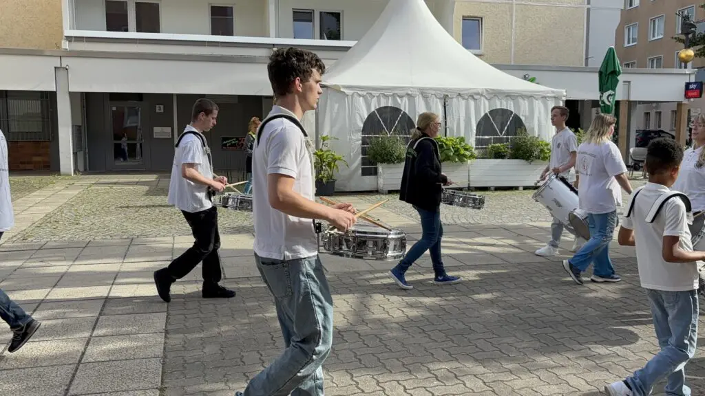 Trommelnde Gruppe in weissen Oberteilen zieht zur Treppenhausmusik 2027 ueber einen gepflasterten Platz vor einem Zelt