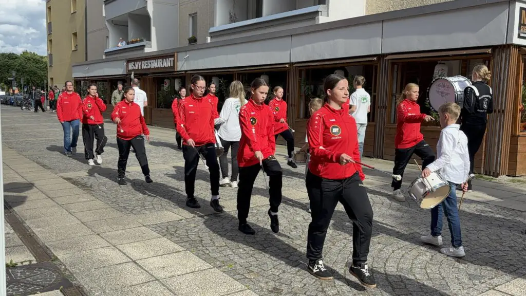 Gruppe in roten Sportjacken marschiert bei Treppenhausmusik 2027 auf dem Gehweg, ein Junge begleitet sie mit einer Trommel