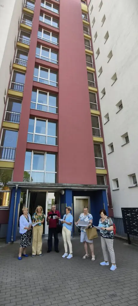 Sechs Personen stehen bei Treppenhausmusik 2027 mit Notenblättern vor einem hohen rot-gelben Wohnhausgebäude