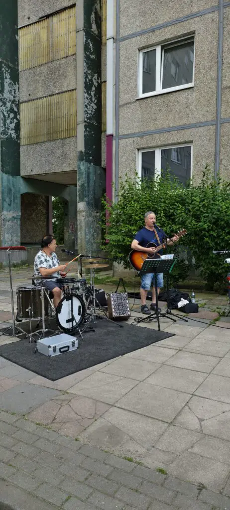 Zwei Musiker spielen bei Treppenhausmusik 2027 mit Gitarre und Schlagzeug vor einem grauen Wohngebäude auf einer Freifläche