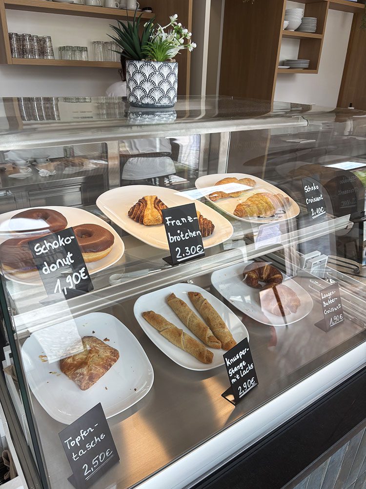 Marienkirchblick-Bistro-Theke: Kuchen, Schoko-Donut, Croissant, Franzbrötchen und Gebäck in der Auslage.