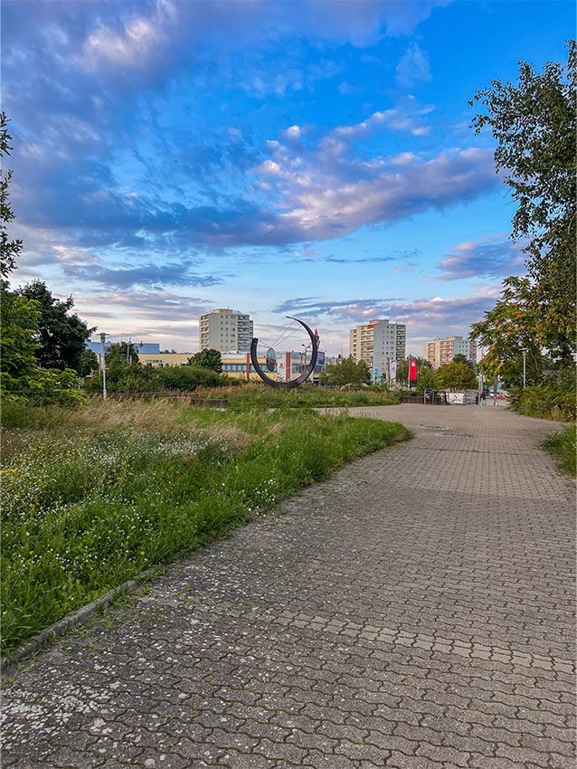 Neuberesinchen Aurorahügel: Blick auf einen gepflasterten Weg mit Kunstobjekt und Wohngebäuden im Hintergrund.