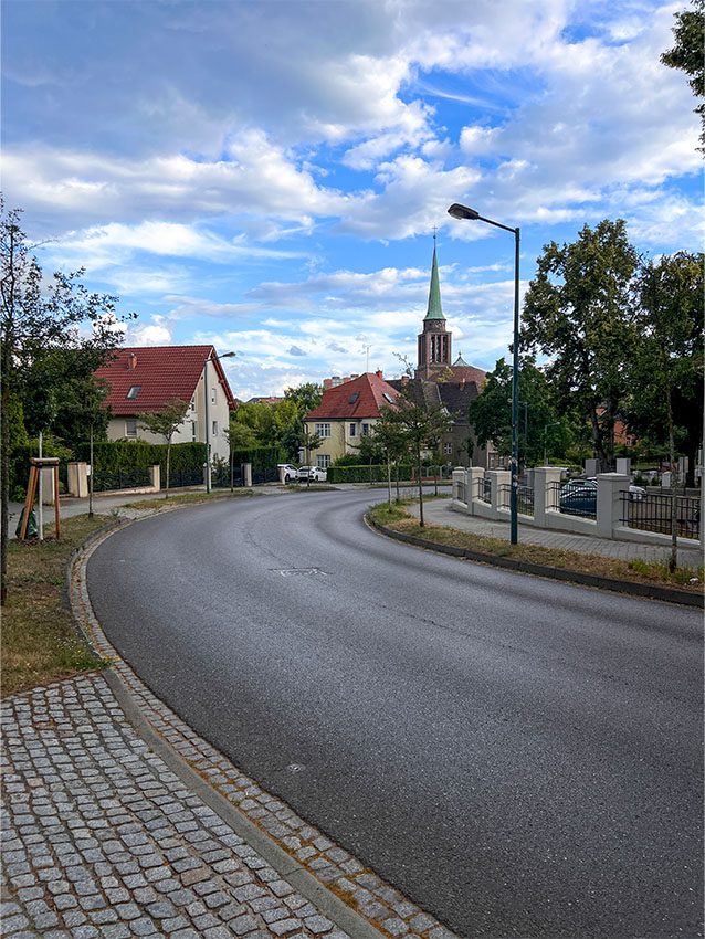 Moskauer Straße: Blick auf eine ruhige Wohnstraße mit Häusern und Kirchturm im Hintergrund.