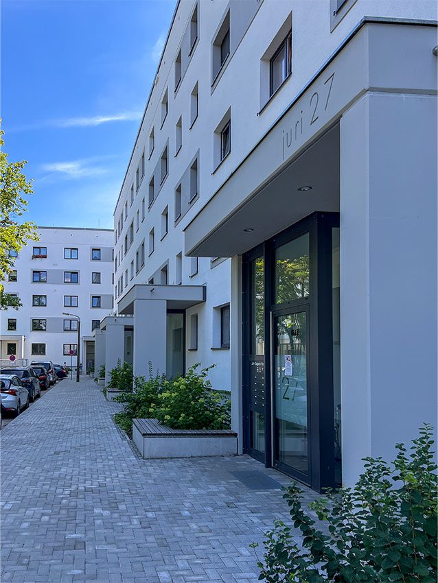 WOWI-FFO-Stadtteil: Fassade eines weißen Mehrfamilienhauses mit Eingangsbereich und Pflasterweg.