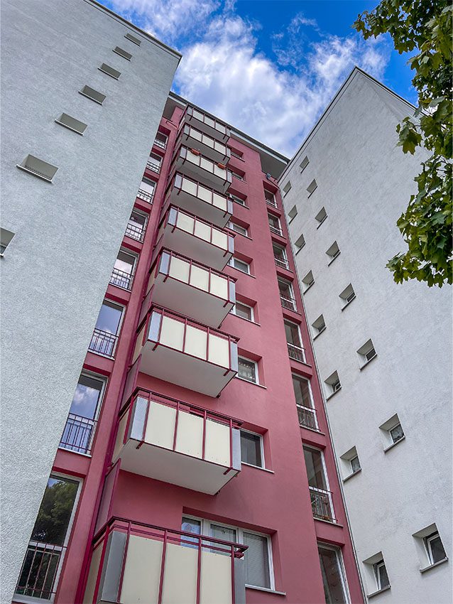 Mehrfamilienhaus mit Balkonen, Fassade in Rosa und Weiß, Blick von unten, blauer Himmel.