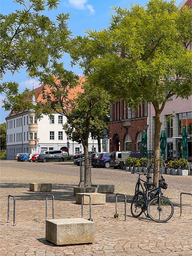 WOWI-FFO-Stadtteil-Zentrum: Platz mit Bäumen, Bänken, Fahrradständern und Gebäuden im Hintergrund. Autos parken.