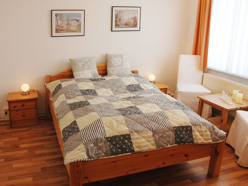 WOWI-FFO-Möbliertes Wohnen: Bett mit Patchworkdecke, Nachttisch, Lampe, Bilder an der Wand, Stuhl, Tisch mit Kerzen.