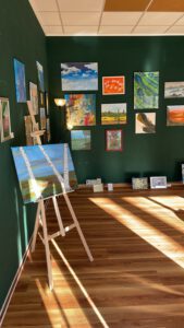 Wander-Atelier der Talente: Gemäldeausstellung mit Birkenmotiv auf Staffelei, umgeben von weiteren Kunstwerken an grüner Wand.