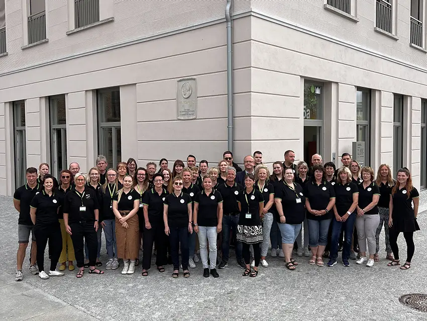 Team-quer Gruppenfoto des Team quer vor einem Gebäude. Die Mitarbeiter tragen einheitliche schwarze Poloshirts mit Logo.