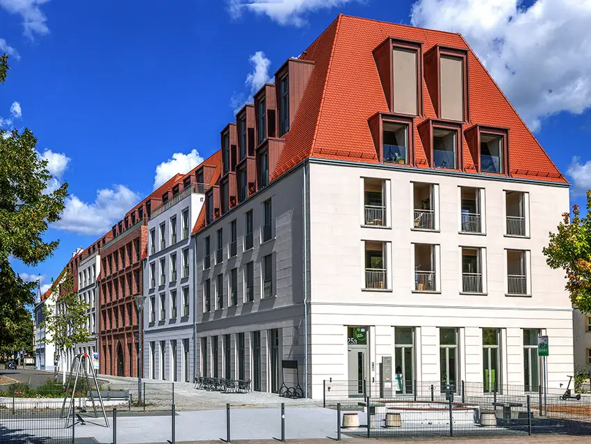 Wie-wir-arbeiten Wie wir arbeiten: Mehrgeschossiges Gebäude mit rotem Dach unter blauem Himmel, Teil einer Häuserreihe, außen Sitzgelegenheiten.