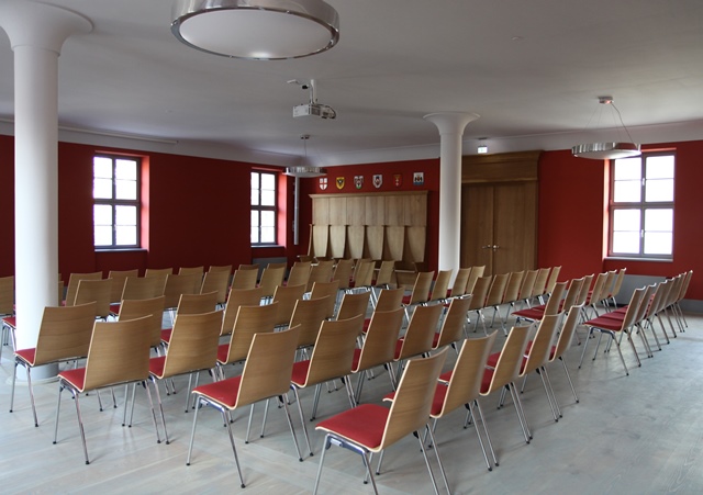 Hansesaal: Innenansicht des Raumes mit Stuhlreihen, Säulen und Wappen an der Wand, geeignet für Veranstaltungen und Tagungen.