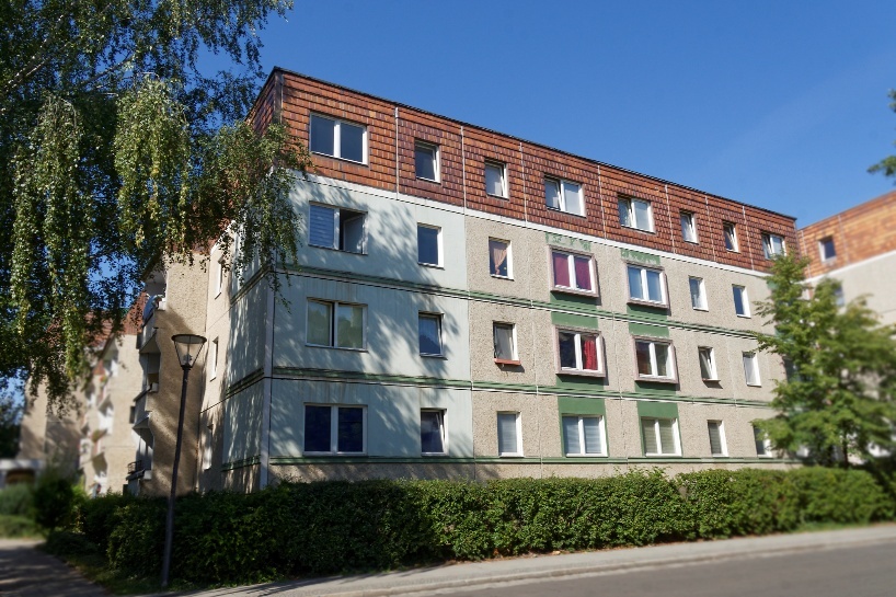 Schulstraße-vor-sanierung Schulstrasse vor Sanierung: Mehrgeschossiges Wohngebäude mit braunem Dach und grünen Akzenten unter blauem Himmel.