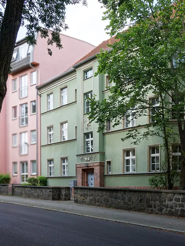 WOWI-FFO-Stadtteil-Altberesinchen: Fassade eines hellgrünen Mietshauses mit weißen Fensterrahmen und einem roten Dach, umgeben von Bäumen.