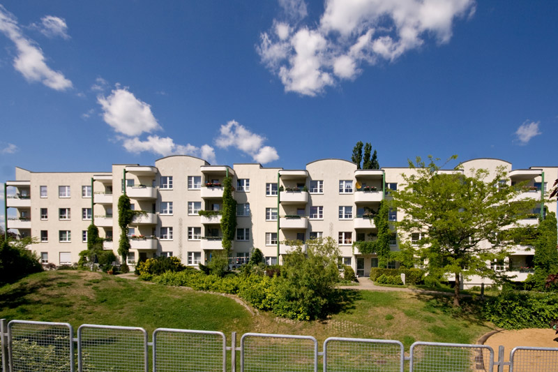 Mieten: Nord_3299 zeigt ein mehrstöckiges Wohngebäude mit Balkonen unter blauem Himmel und Wolken, umgeben von Grünflächen.